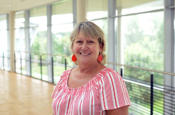 Porträtfoto: Jutta Meyborn im audimax. Sie trägt ein rot-weiß gestreiftes Oberteil und große, rote Ohrringe. Hinter ihr ist die Fensterfront des audimax mit Ausblick auf den grünen Campus und der Parkettboden zu sehen.