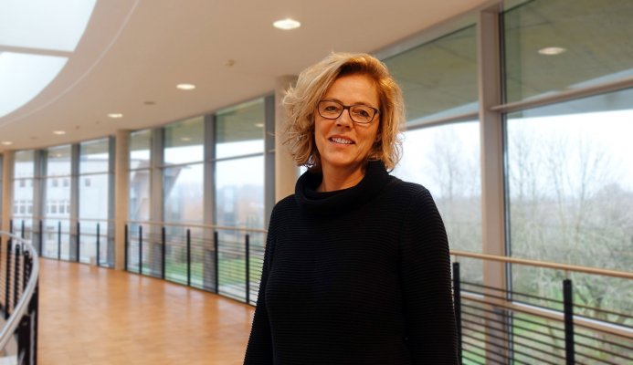 Porträtfoto: Maja Petersen im audimax. Sie trägt ein dunkles Oberteil und Brille. Wellige blonde Haare reichen bis zum Kinn. Hinter ihr ist die Fensterfront des audimax mit Ausblick auf den grünen Campus und der Parkettboden zu sehen.