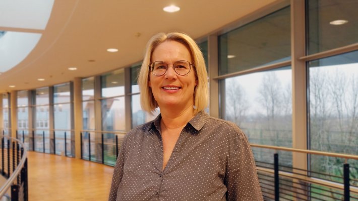 Porträtfoto: Manuela Petersen im audimax. Sie trägt eine graue Bluse und Brille. Ein blonder Bob reicht bis zum Kinn. Hinter ihr ist die Fensterfront des audimax mit Ausblick auf den grünen Campus und der Parkettboden zu sehen.