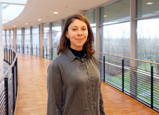 Porträtfoto: Rebekka Küchler im audimax. Sie trägt ein graues Oberteil. Ihre Haare sind dunkel und etwa schulterlang, sie trägt sie offen. Hinter ihr ist die Fensterfront des audimax mit Ausblick auf den grünen Campus und der Parkettboden zu sehen.