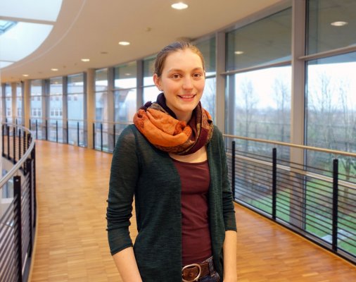 Porträtfoto: Samanta Rohm im audimax. Sie trägt ein weinrotes Oberteil, darüber einen olivgrünen Cardigan und einen Schal mit kräftigen Farben. Ihre Haare trägt sie zusammen gebunden. Hinter ihr ist die Fensterfront des audimax mit Ausblick auf den grünen Campus und der Parkettboden zu sehen.