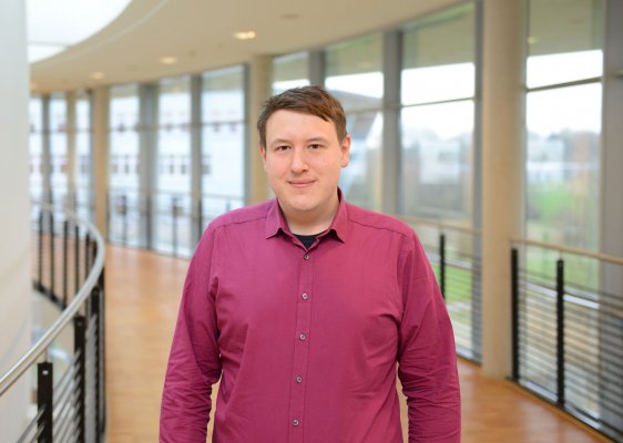 Portraitfoto: Johann Kirchkneser im audimax. Er trägt ein beerenfarbenes Hemd. Seine Haare sind kurz. Hinter ihm ist die Fensterfront des audimax mit Ausblick auf den grünen Campus und der Parkettboden zu sehen.