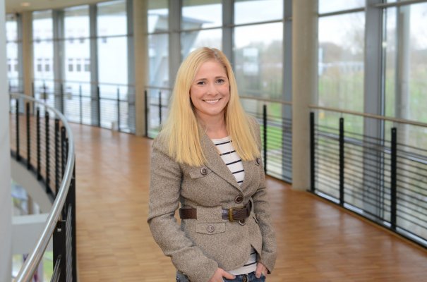 Porträtfoto: Nelly Oelze im audimax. Sie trägt einen grauen Blazer über einem  gestreiften  Oberteil. Ihre blonden Haare trägt sie offen. Hinter ihr ist die Fensterfront des audimax mit Ausblick auf den grünen Campus und der Parkettboden zu sehen.