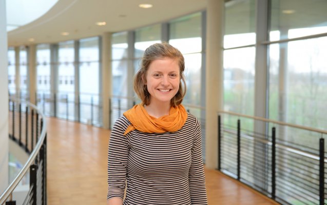 Portraitfoto: Carina Schmidt im audimax. Sie trägt ein graues Oberteil und einen Schald in einem gelblichen orange. Ihre dunkelblonden etwa schulterlangen Haare sind locker zurückgebunden. Sie lächelt. Hinter ihr ist die Fensterfront des audimax mit Ausblick auf den grünen Campus und der Parkettboden zu sehen.