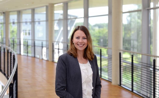 Porträtfoto: Marlena Baron im audimax. Sie trägt einen dunklen Blazer über einer weißen Bluse. Ihre dunkelblonden Haare trägt sie offen. Hinter ihr ist die Fensterfront des audimax mit Ausblick auf den grünen Campus und der Parkettboden zu sehen.