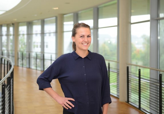 Porträtfoto: Lisa-Marie Cig im audimax. Sie trägt eine blaue Bluse, die Haare sind zusammengebunden und die rechte Hand in der Hüfte. Hinter ihr ist die Fensterfront des audimax mit Ausblick auf den grünen Campus und der Parkettboden zu sehen.