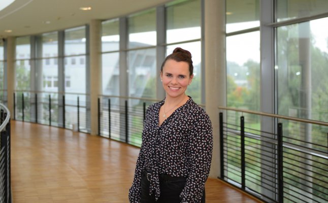 Porträtfoto: Stefanie Jordt im audimax. Sie trägt eine gemusterte Bluse, die Haare im Dutt. Hinter ihr ist die Fensterfront des audimax mit Ausblick auf den grünen Campus und der Parkettboden zu sehen.