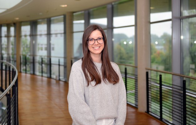 Porträtfoto: Beate Panitz im audimax. Sie trägt einen hellen Pullover über einer weißen Bluse, die langen Haare trägt sie offen. Hinter ihr ist die Fensterfront des audimax mit Ausblick auf den grünen Campus und der Parkettboden zu sehen.