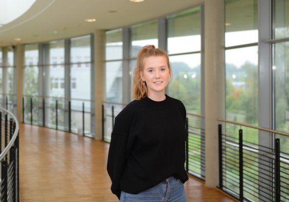 Porträtfoto: Julia Redepennig im audimax. Sie trägt einen dunklen Pullover und Jeans, die Haare im Pferdeschwanz. Hinter ihr ist die Fensterfront des audimax mit Ausblick auf den grünen Campus und der Parkettboden zu sehen.