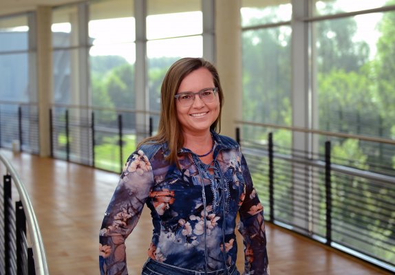 Porträtfoto: Kim Beisel im audimax. Sie trägt ein gemustertes Oberteil und Jeans. Hinter ihr ist die Fensterfront des audimax mit Ausblick auf den grünen Campus und der Parkettboden zu sehen.