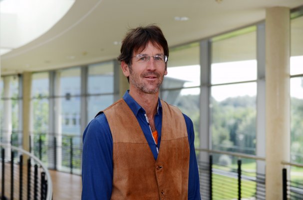 Porträtfoto: Christian Brandt im audimax. Er trägt ein blaues Hemd mit brauner Weste. Hinter ihm ist die Fensterfront des audimax mit Ausblick auf den grünen Campus und der Parkettboden zu sehen.