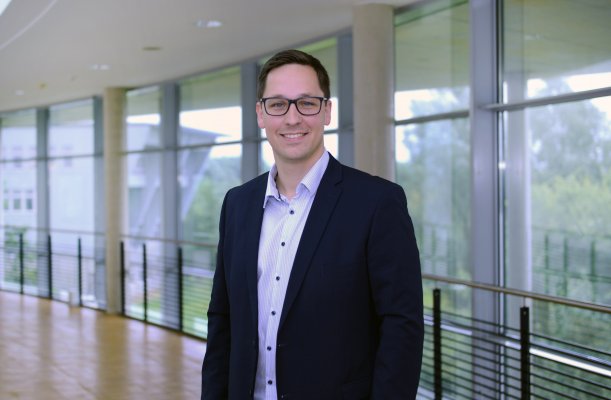 Porträtfoto: Leo Eriksen im audimax. Er trägt ein helles Hemd mit dunklem Jackett. Hinter ihm ist die Fensterfront des audimax mit Ausblick auf den grünen Campus und der Parkettboden zu sehen.