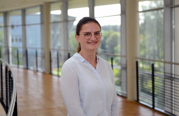 Porträtfoto: Ronja Isert im audimax. Sie trägt eine weiße Bluse mit hellblauen Streifen. Hinter ihr ist die Fensterfront des audimax mit Ausblick auf den grünen Campus und der Parkettboden zu sehen.