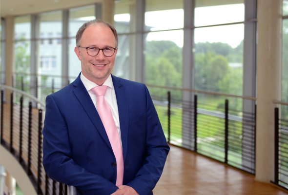 Porträtfoto: Christoph Jansen im audimax. Er trägt ein weißes Hemd und dunkelblaues Jackett. Hinter ihm ist die Fensterfront des audimax mit Ausblick auf den grünen Campus und der Parkettboden zu sehen.