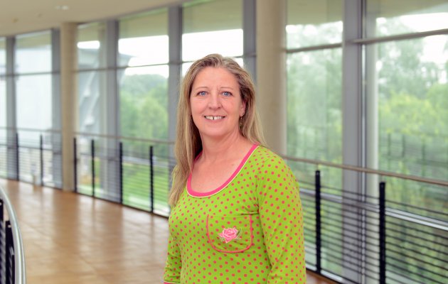 Porträtfoto: Iris Mainusch im audimax. Sie trägt ein grünes Oberteil mit pink-farbenen Punkten. Hinter ihr ist die Fensterfront des audimax mit Ausblick auf den grünen Campus und der Parkettboden zu sehen.