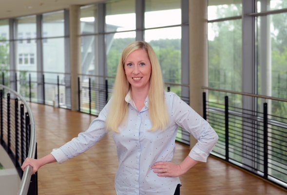 Porträtfoto: Kimmy Ormstrup im audimax. Sie trägt eine weiße Bluse. Hinter ihr ist die Fensterfront des audimax mit Ausblick auf den grünen Campus und der Parkettboden zu sehen.