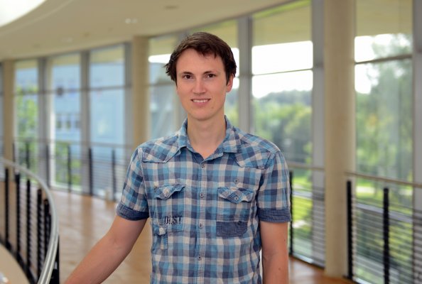 Porträtfoto: Christoph Thomsen im audimax. Er trägt ein blau-eweiß-kariertes Hemd. Hinter ihm ist die Fensterfront des audimax mit Ausblick auf den grünen Campus und der Parkettboden zu sehen.