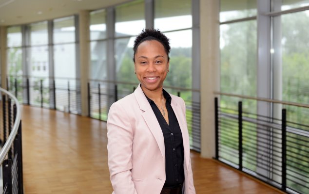 Porträtfoto: Aadila Cayenne im audimax. Sie trägt einen hellen Blazer über einem dunklen Oberteil. Hinter ihr ist die Fensterfront des audimax mit Ausblick auf den grünen Campus und der Parkettboden zu sehen.