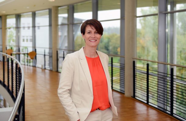 Porträtfoto: Silke Griemsmann im audimax. Sie trägt kurze dunkle Haare und ein orange-farbenes Oberteil unter einem hellen Blazer. Hinter ihr ist die Fensterfront des audimax mit Ausblick auf den grünen Campus und der Parkettboden zu sehen.