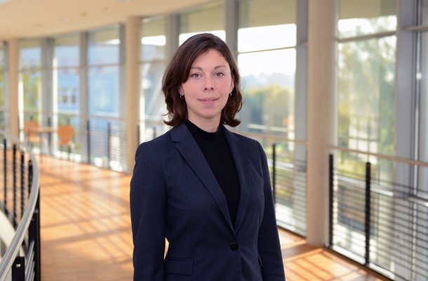 Porträtfoto: Rebekka Küchler im audimax. Sie trägt ein dunkles Oberteil unter einem dunklen Blazer, die dunklen Haare sind schulterlang. Ihre Haare sind dunkel und etwa schulterlang, sie trägt sie offen. Hinter ihr ist die Fensterfront des audimax mit Ausblick auf den grünen Campus und der Parkettboden zu sehen.