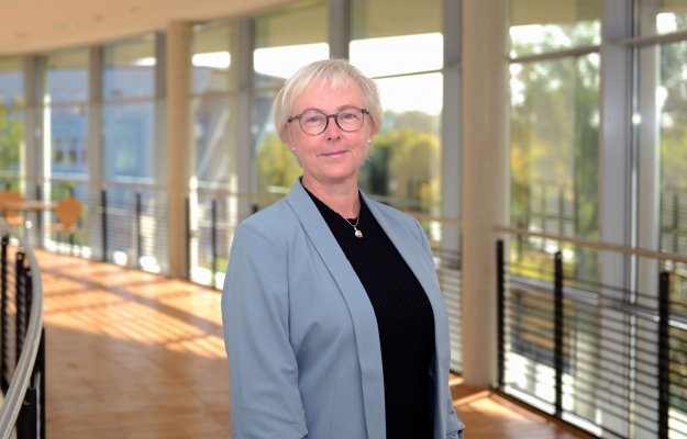 Porträtfoto: Monika Tinzmann im audimax. Sie trägt ein dunkles Oberteil unter einem grauen Blazer, Brille und kurze blonde Haare. Hinter ihr ist die Fensterfront des audimax mit Ausblick auf den grünen Campus und der Parkettboden zu sehen.