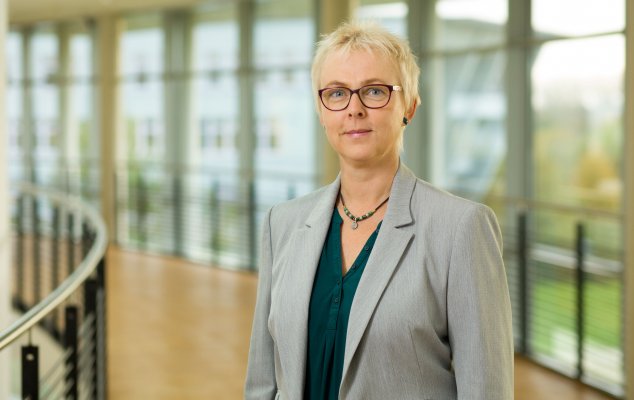 Porträtfoto: Monika Tinzmann im audimax. Sie trägt eine dunkelgrüne Bluse unter einem grauen Blazer, Brille und kurze blonde Haare. Hinter ihr ist die Fensterfront des audimax mit Ausblick auf den grünen Campus und der Parkettboden zu sehen.