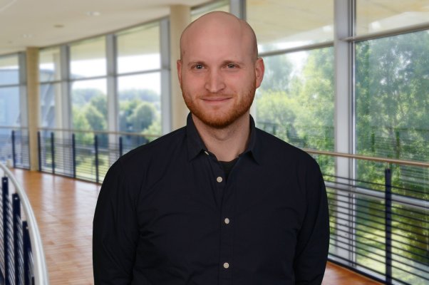 Porträtfoto: Henner Bendig im audimax. Er trägt ein dunkles Hemd, einen leichten Bart und Glatze. Hinter ihm ist die Fensterfront des audimax mit Ausblick auf den grünen Campus und der Parkettboden zu sehen.