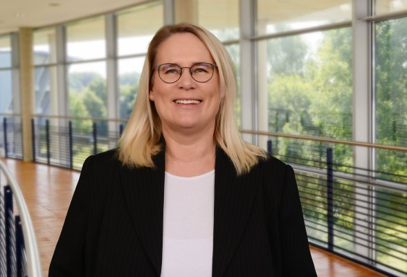 Porträtfoto: Manuela Petersen im audimax. Sie trägt einen dunklen Blazer über einem helleb Oberteil und Brille. Die blonden Haare reichen über die Schulter. Hinter ihr ist die Fensterfront des audimax mit Ausblick auf den grünen Campus und der Parkettboden zu sehen.