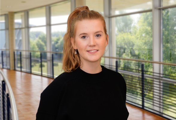 Porträtfoto: Julia Redepennig im audimax. Sie trägt einen dunklen Pullover und Jeans, die Haare im Pferdeschwanz. Hinter ihr ist die Fensterfront des audimax mit Ausblick auf den grünen Campus und der Parkettboden zu sehen.