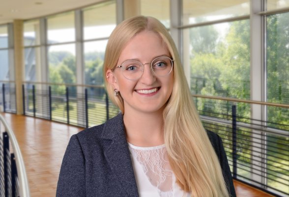 Porträtfoto: Britta Blotenberg im audimax, sie trägt die langen, blonden Haare offen über die linke Schulter und einen dunklen Blazer über einem weißen Oberteil.