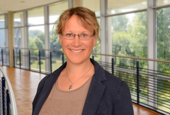 Portraitfoto von Johanna Siebel-Mogk. Sie steht im Audimax der Hochschule Flensburg, trägt eine Brille und die blonden Haare zusammengebunden. Sie trägt einen dunkelblauen Blazer über einem hellen Oberteil.