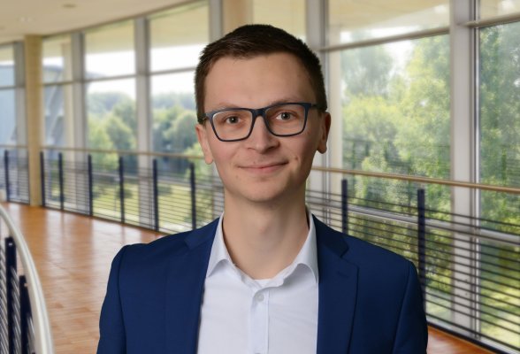 Porträtfoto: Carl Hendrik Mennerich im audimax, er trägt kurze dunkelblonde Haare und eine Brille. Über einem hellen Hemd trägt er ein dunkelblaues Jackett.