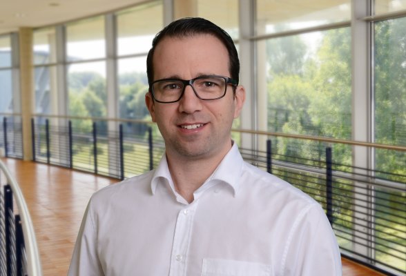 Porträtfoto: Andreas Heinen im audimax, er trägt kurze dunkle Haare und ein Brille. Er trägt ein weißes Hemd.