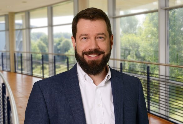 Porträtfoto: Niklas Klein im audimax, er trägt die dunklen Haaare kurz, dazu einen Bart und ein dunkles Jackett über einem weißen Hemd.