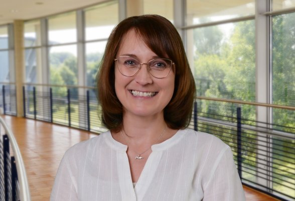 Porträtfoto: Manuela Schallenburger im audimax, sie trägt die schulterlangen, dunklen Haare offen, eine Brille und eine weiße Bluse.