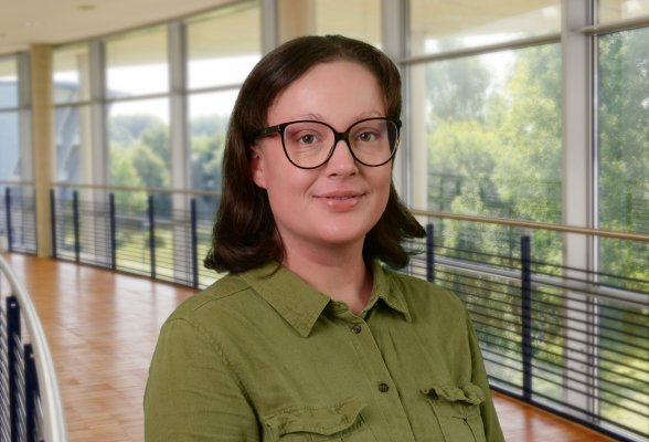 Porträtfoto: Rebecca Starke im audimax, sie trägt die schulterlangen, dunklen Haare offen, eine Brille und eine grüne Bluse.