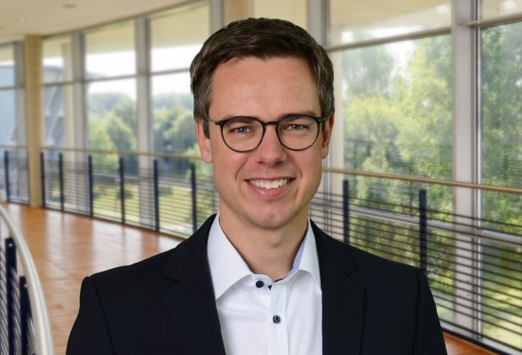 Porträtfoto: Lasse Tausch-Nebel im audimax, er trägt dunkle, kurze Haaare, eine Brille und ein dunkles Jackett über einem weißen Hemd.