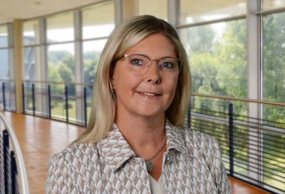 Porträtfoto: Katja Roberts im audimax, sie trägt die schulterlangen, blonden Haare offen, eine Brille und eine helle, gemusterte Jacke über einem beige-farbenen Oberteil