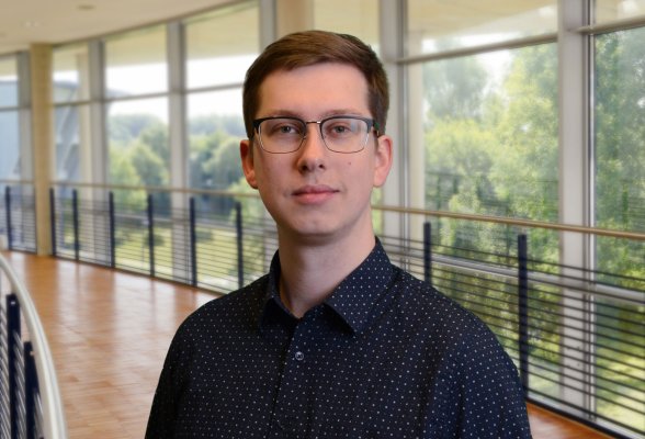 Porträtfoto: Pascal Friedrichsen im audimax, er trägt dunkelblonde, kurze Haaare, eine Brille und ein dunkles Hemd mit kleinen hellen Punkten.