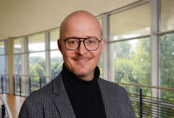 Portraitfoto: Bosco Lehr im audimax, er trägt Glatze und einen leichten Bart; einen dunklen Rollkragenpullover unter einem grau-schwarz karierten Jackett und eine Brille mit dunklem Rahmen.