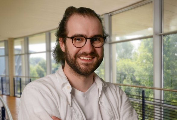 Porträtfoto: Tobis Friedel im audimax, seine Haare sind dunkelblond, lockig und zu einem Dutt gebunden. Er trägt Bart, eine Brille und ein weißes Hemd über einem weißen Shirt.