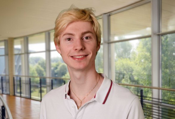 Porträtfoto: Reinhard Jäkel im audimax, er trägt blonde, kurze Haare und ein weißes Polohemd. Um den Hals trägt er eine goldene Kette.