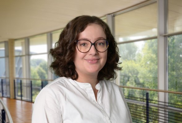 Porträtfoto: Rebecca Starke im audimax, sie trägt die schulterlangen, dunklen Locken offen, eine Brille und eine weiße Bluse.