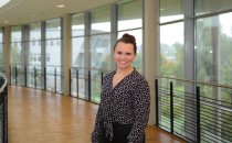 Porträtfoto: Stefanie Jordt im audimax. Sie trägt eine gemusterte Bluse, die Haare im Dutt. Hinter ihr ist die Fensterfront des audimax mit Ausblick auf den grünen Campus und der Parkettboden zu sehen.