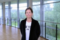 Porträtfoto: Maika Worm im audimax. Sie trägt ein weißes Oberteil und einen dunklen Blazer. Hinter ihr ist die Fensterfront des audimax mit Ausblick auf den grünen Campus und der Parkettboden zu sehen.