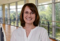 Porträtfoto: Manuela Schallenburger im audimax, sie trägt die schulterlangen, dunklen Haare offen, eine Brille und eine weiße Bluse.
