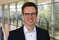 Porträtfoto: Lasse Tausch-Nebel im audimax, er trägt dunkle, kurze Haaare, eine Brille und ein dunkles Jackett über einem weißen Hemd.