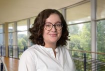 Porträtfoto: Rebecca Starke im audimax, sie trägt die schulterlangen, dunklen Locken offen, eine Brille und eine weiße Bluse.