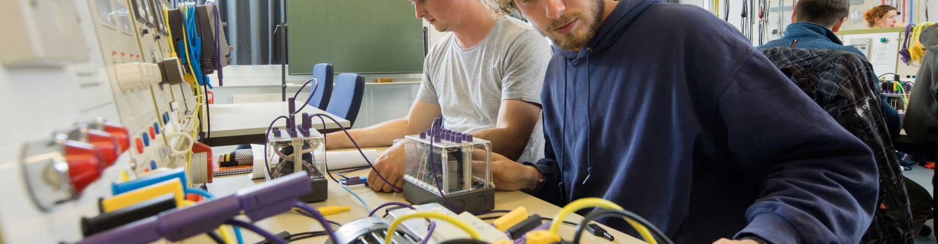 Zwei Studenten arbeiten an einer elektrotechnischen Maschine, zu sehen sind Kabel in unterschiedlichen Fraben und rote Leuchten. Im Hintergrund eine Tafel und weitere Studierende, die an ähnlichen Versuchen arbeiten.
