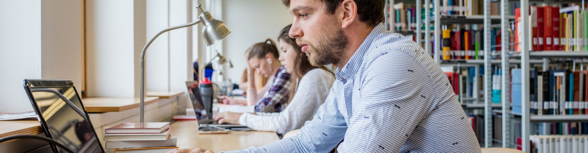 Eine Bibliothek. Rechts sind Bücherregale zu erkennen, links arbeiten mehrere Studierende an einem langen Tisch. Vorn im Bild ein kurzhaariger Mann im blauen Hemd, der an einem Laptop arbeitet.